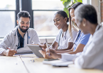 A small group of four medical professionals sit around a boardroom table as they meet to discuss patient cases. They are each dressed professionally in scrubs and lab coats as they focus on working together.