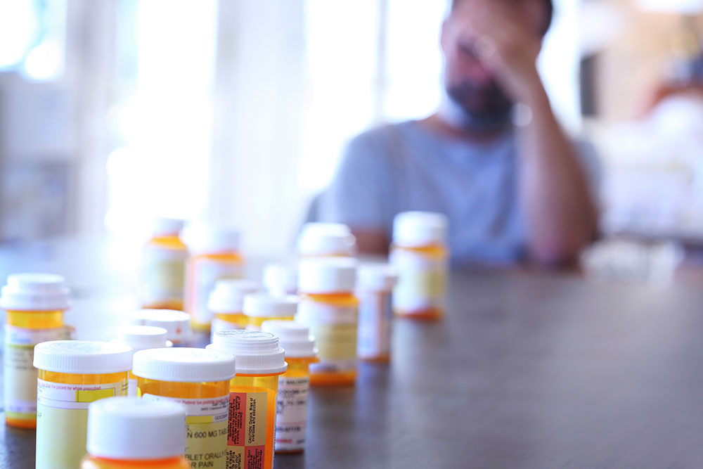 A large group of prescription medication bottles sit on a table as a man in the background sits at the end of the table with his hand on his head.. The image is photographed with a very shallow depth of field with the focus being on the pill bottles in the foreground.