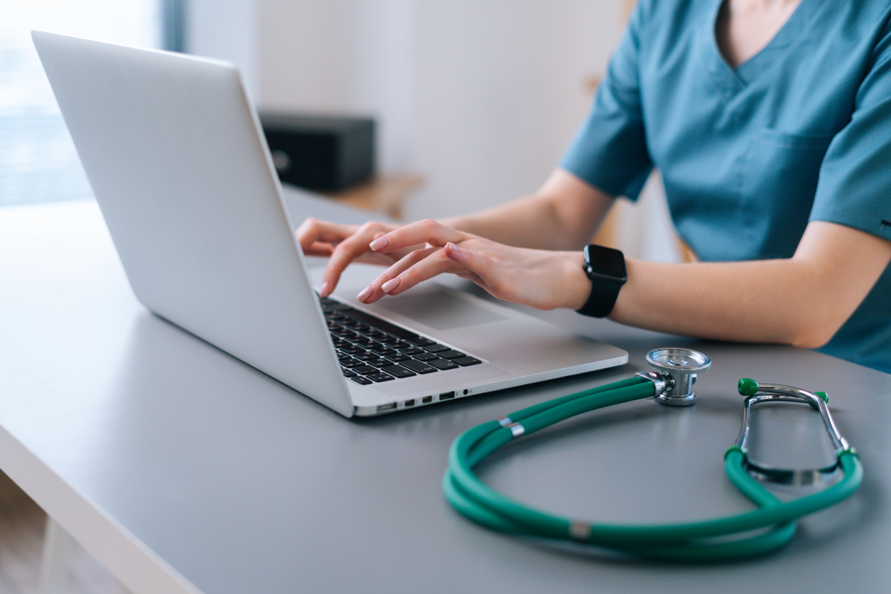 Close-up hands of unrecognizable female physician in medical uniform working typing on laptop keyboard sitting at desk on background of window in dark office room