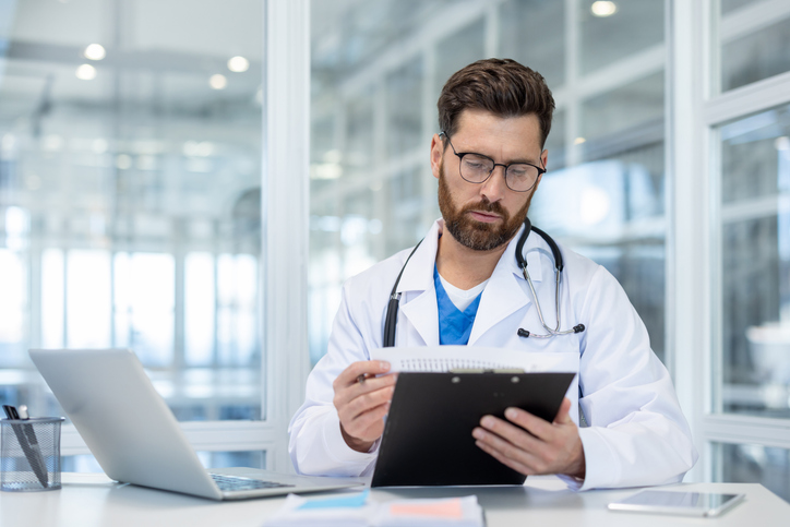 A doctor in a white coat reviews medical records, with a laptop and pen holder visible on the desk. The healthcare professional is wearing glasses.
